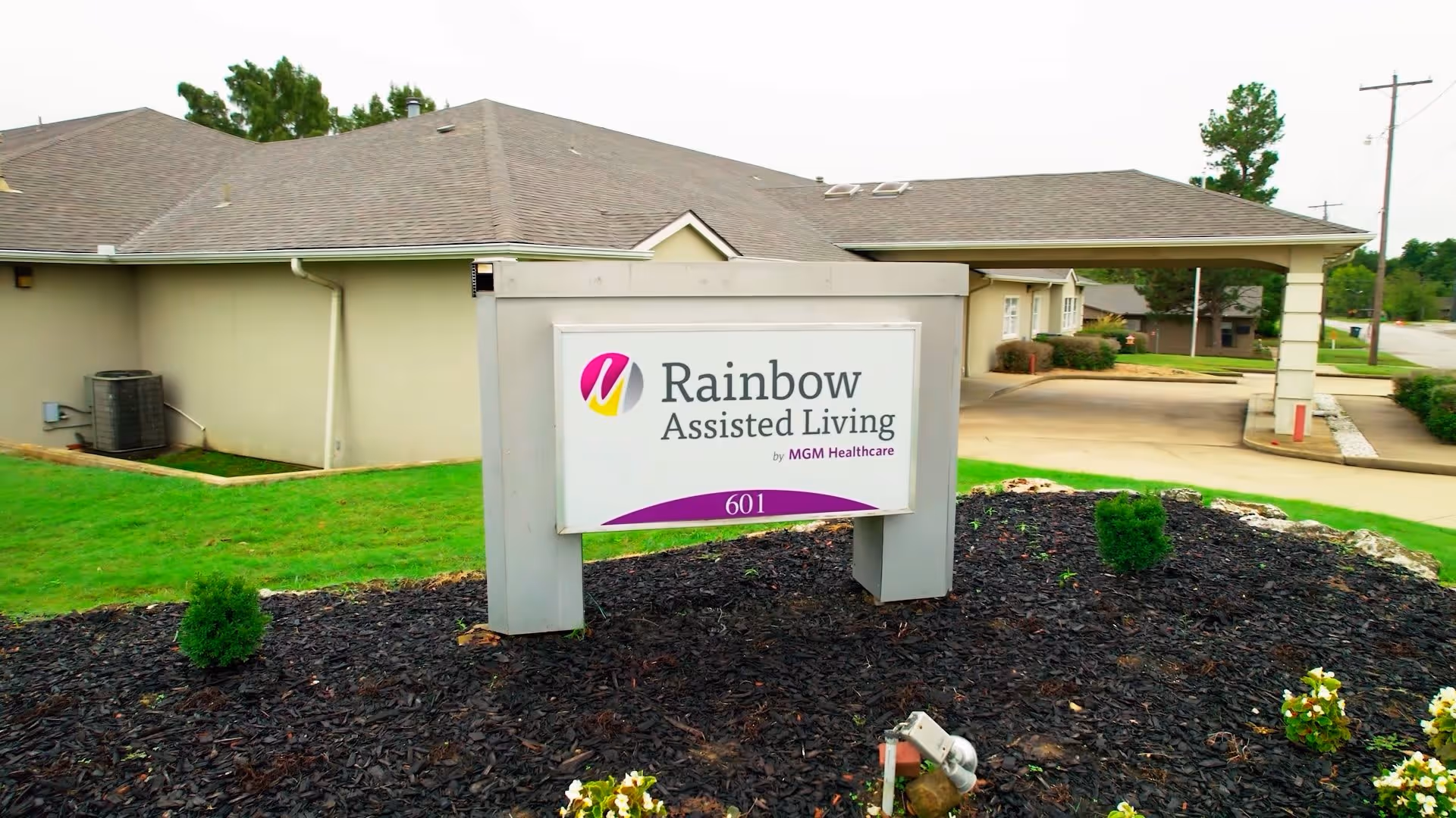 A freestanding sign reading "Rainbow Assisted Living" stands in a landscaped bed in front of a single-story facility and drive-through entrance.