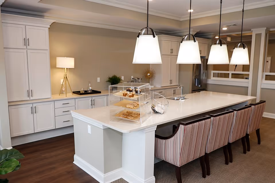 A modern kitchen area with a large white island countertop featuring a sink and four striped upholstered chairs. Above the island, there are four pendant lights. On the countertop, there is a clear display case with pastries and cookies. The background shows white cabinetry, a lamp on the counter, and a stainless steel refrigerator.