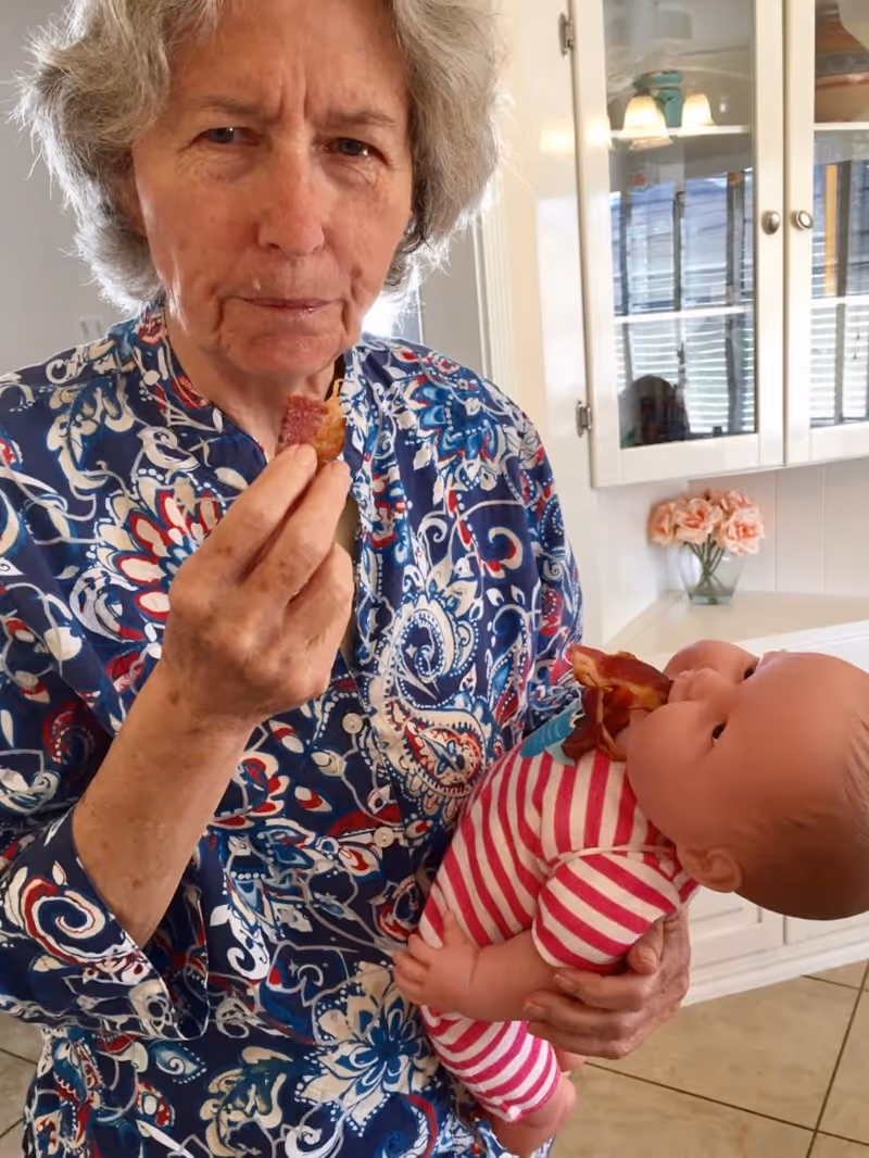 An elderly woman wearing a blue patterned shirt is holding a baby doll dressed in pink and white striped clothing. Both the woman and the doll have pieces of bacon near their mouths. The background shows a white kitchen cabinet with glass doors and a vase with pink flowers on the counter.
