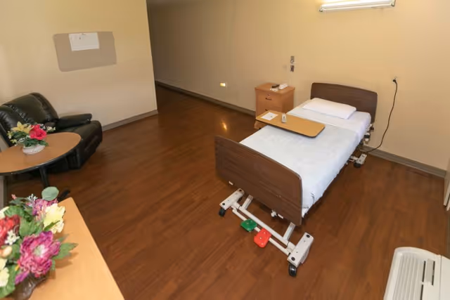 A simple room with a single hospital bed equipped with a white pillow and a tray table. Next to the bed is a wooden nightstand with a telephone and a small sign. The room has wooden flooring, beige walls, a black leather armchair with a round table holding a flower arrangement, and a heating or air conditioning unit near the floor.