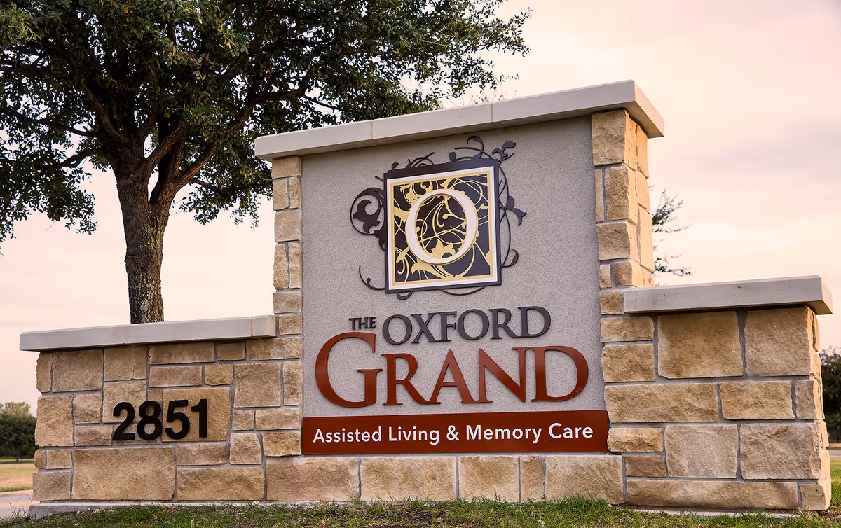 Stone and concrete sign for The Oxford Grand Assisted Living & Memory Care with the address number 2851, set outdoors with a tree and a sunset sky in the background.