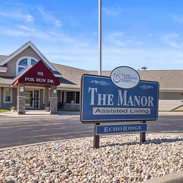 Exterior view of Good Samaritan Society - Echo Ridge - The Manor assisted living facility showing the building entrance with a red awning labeled 931 Fox Run Dr. and a large blue sign in the foreground that reads The Manor Assisted Living Echo Ridge under a clear blue sky.