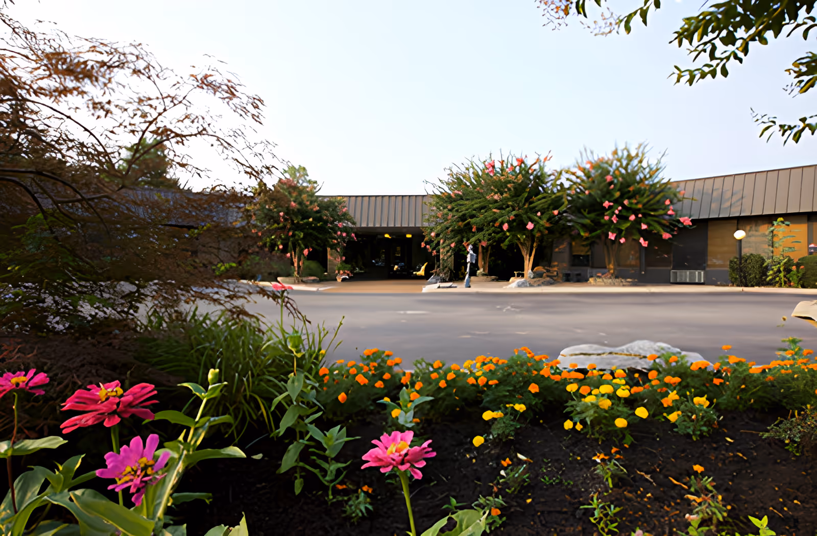 View of the front exterior of a single-story building with a flat roof, surrounded by landscaped flower beds with pink and orange flowers and several small trees with pink blossoms. A person is standing near the entrance of the building.