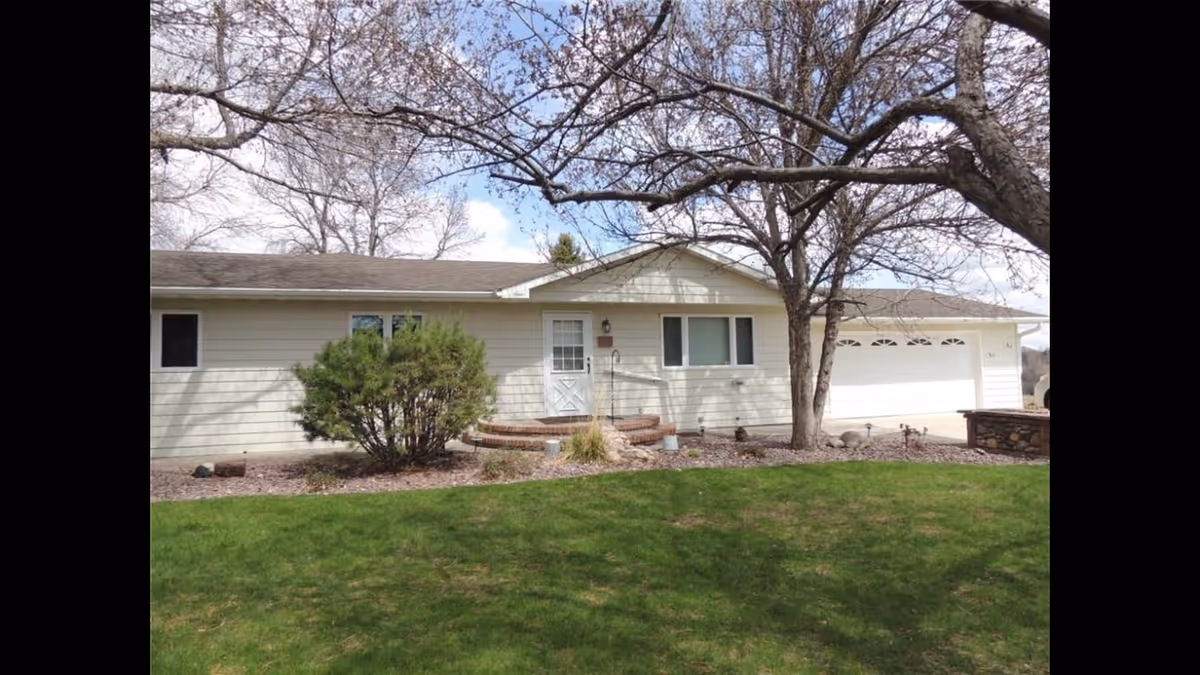 Single-story house with light-colored siding, a white front door, two windows, a two-car garage, and a green lawn in front. There are leafless trees and a bush near the entrance under a partly cloudy sky.