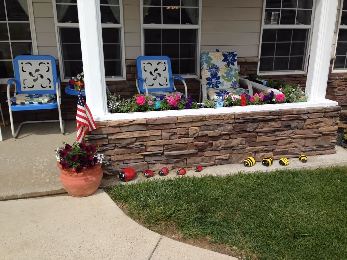 Outdoor patio area with two blue metal chairs and one cushioned chair with floral patterns. A stone planter box filled with colorful flowers is in front of the chairs. A small American flag is placed in a flower pot with dark red flowers on the concrete patio. Decorative painted rocks resembling ladybugs and bees are lined up on the ground near the grass.