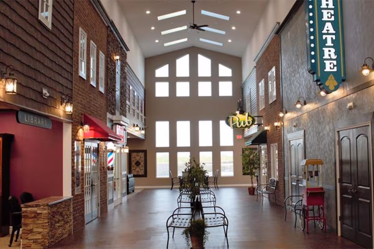 Interior view of a senior living facility hallway designed like a small town street with storefronts labeled Library, Pub, and Theatre. The hallway has high ceilings with skylights, large windows at the end, benches with plants in the center, and warm lighting fixtures on the walls.