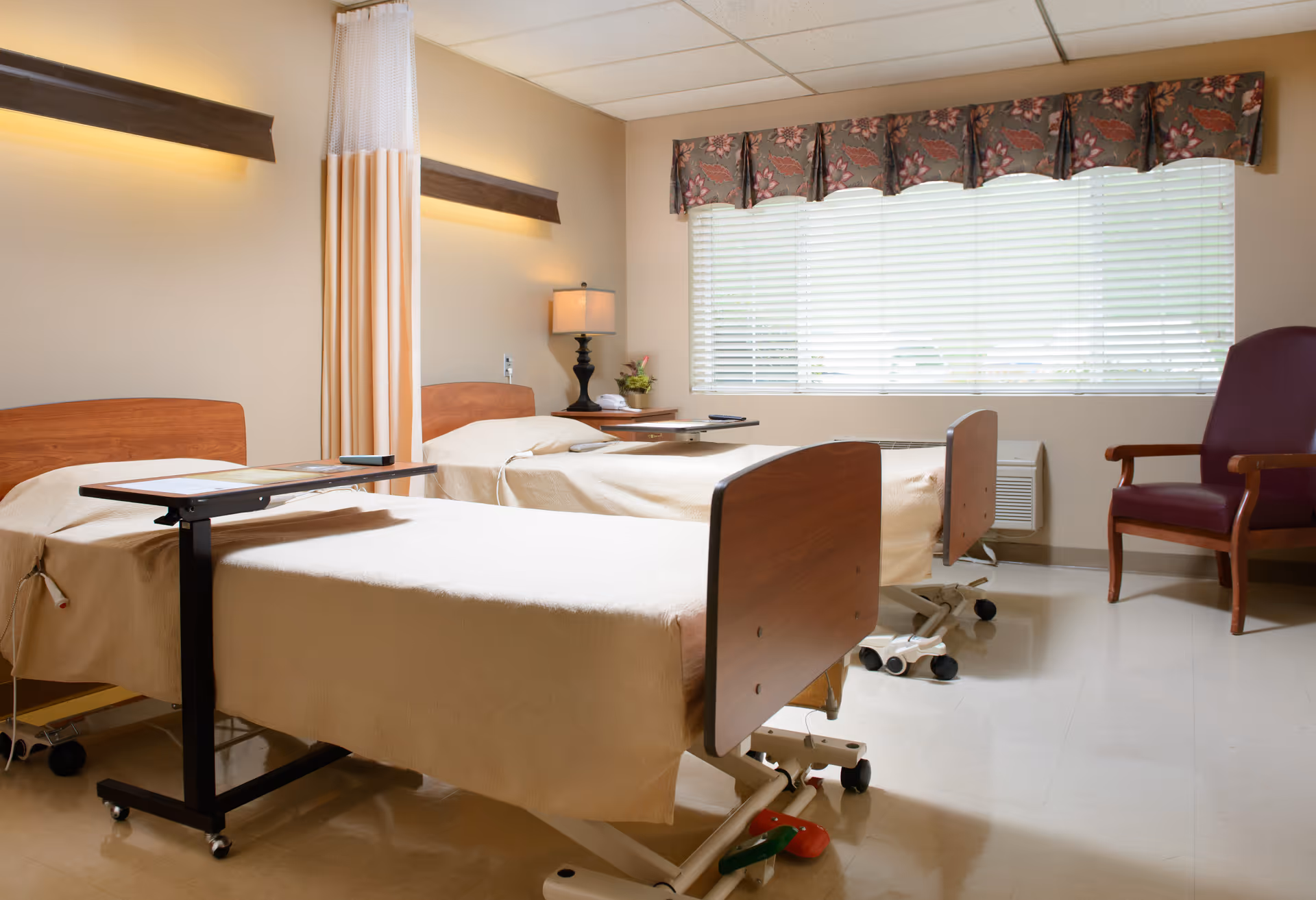 A room with two hospital-style beds covered with beige sheets, each with an overbed table. There is a curtain divider between the beds, a window with blinds and floral valance, a wooden side table with a lamp and a small plant, and a maroon armchair in the corner.