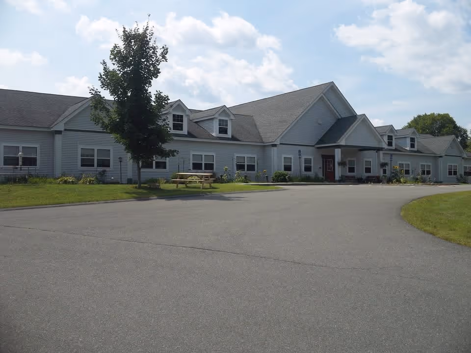 Exterior view of a single-story building with gray siding and a gabled roof under a partly cloudy sky. There is a paved driveway in front, a small tree, and a wooden picnic table on the grass near the building.