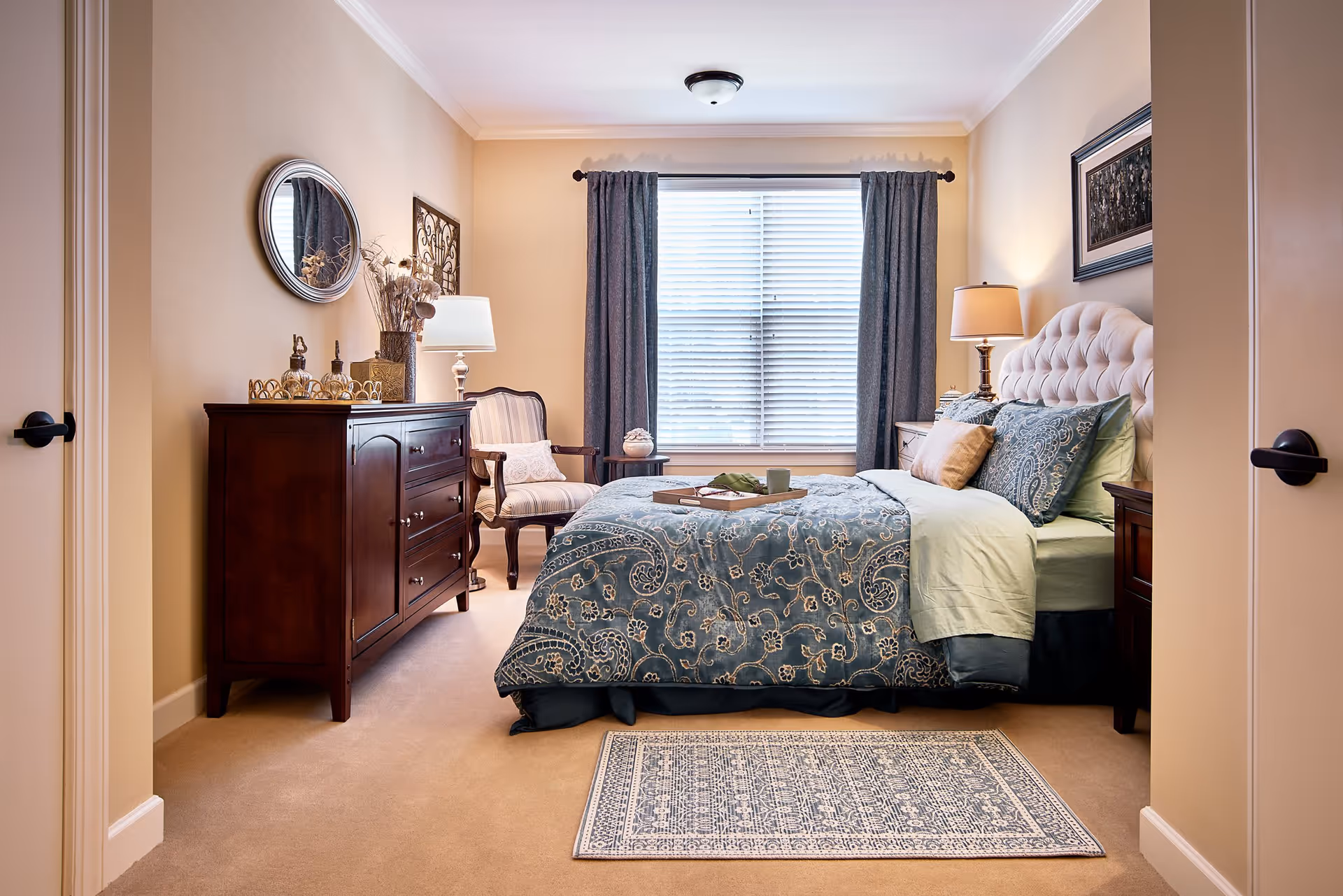 A cozy bedroom with a large bed featuring a blue patterned comforter and multiple pillows. There is a wooden dresser with decorative items and a round mirror on the left wall. A striped armchair with a cushion is next to the dresser. The room has beige walls, a window with closed blinds and blue curtains, two bedside tables with lamps, and a patterned rug on the floor.