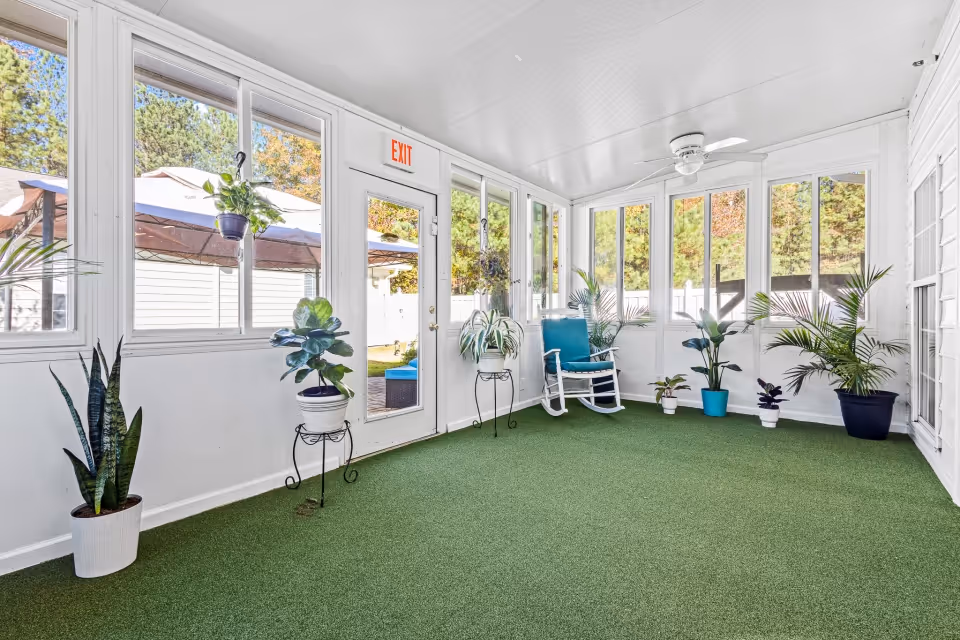 Bright enclosed sunroom with green carpeting, potted plants, a blue rocking chair, and many windows.