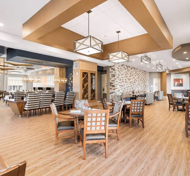A spacious and well-lit dining area with wooden floors, multiple tables and chairs arranged for seating, modern pendant lights hanging from the ceiling, and a decorative wall with a mosaic tile pattern. The room has a warm and inviting atmosphere with a mix of patterned and striped upholstery on the chairs.