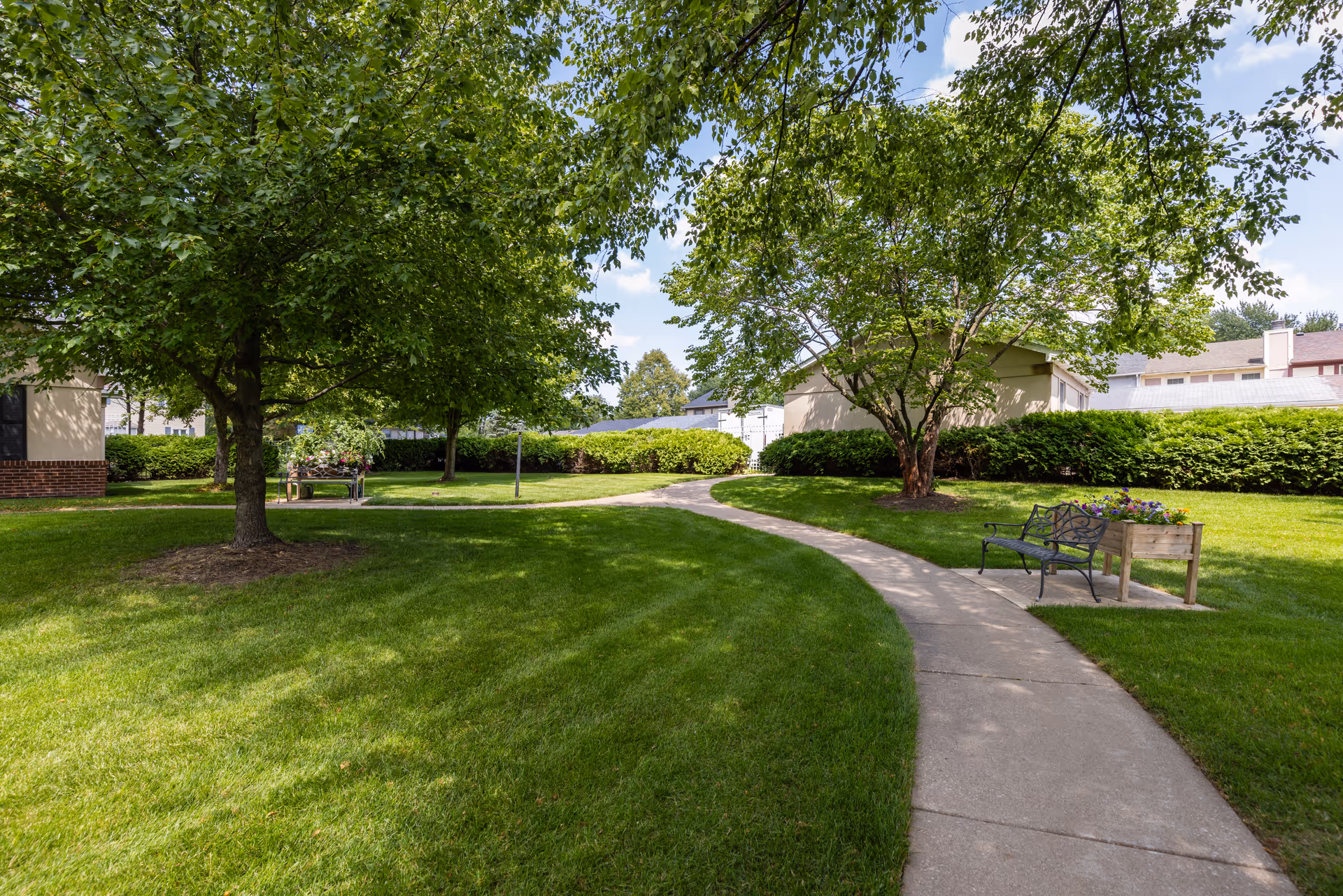 A curved concrete pathway winds through a well-maintained green lawn with trees providing shade. There are benches and flower planters along the path, with buildings and hedges visible in the background under a partly cloudy sky.