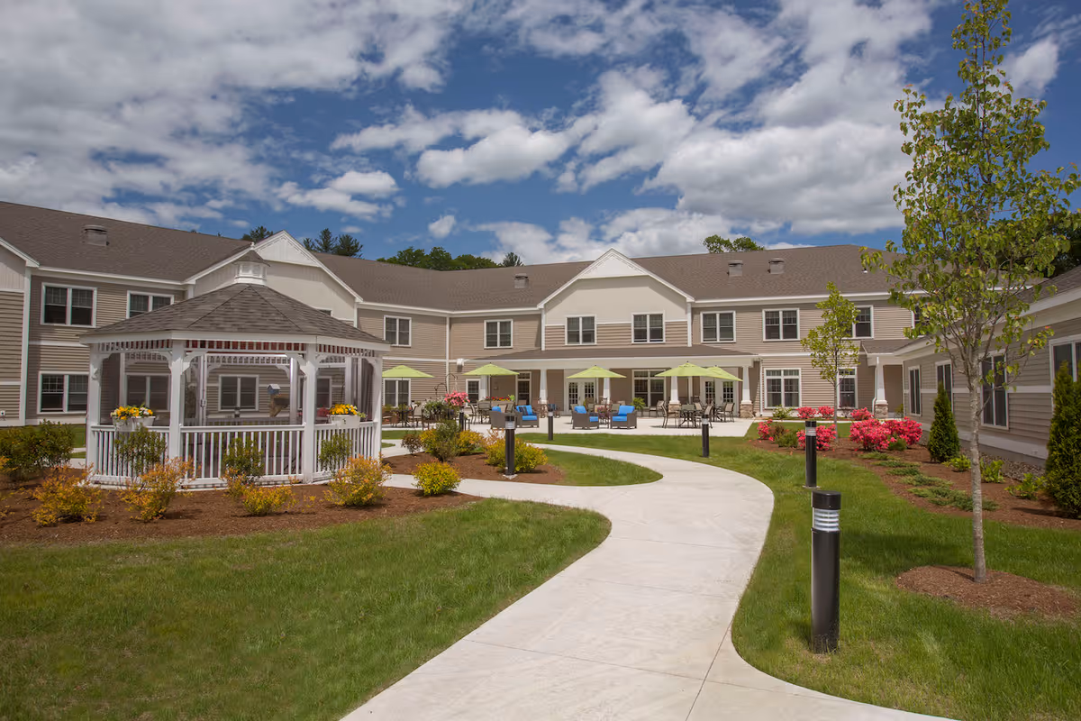 Outdoor courtyard area of a senior living facility with a winding concrete pathway, green grass, small trees, and flower beds. There is a white gazebo on the left side and several patio tables with green umbrellas and blue chairs near the building. The building is two stories with beige siding and white trim under a partly cloudy blue sky.