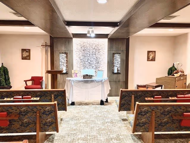 Interior view of a small chapel or prayer room with wooden pews facing an altar covered with a white cloth. The altar has religious items on it, and there is a cross on the left wall. The room has soft lighting and religious artwork on the walls.
