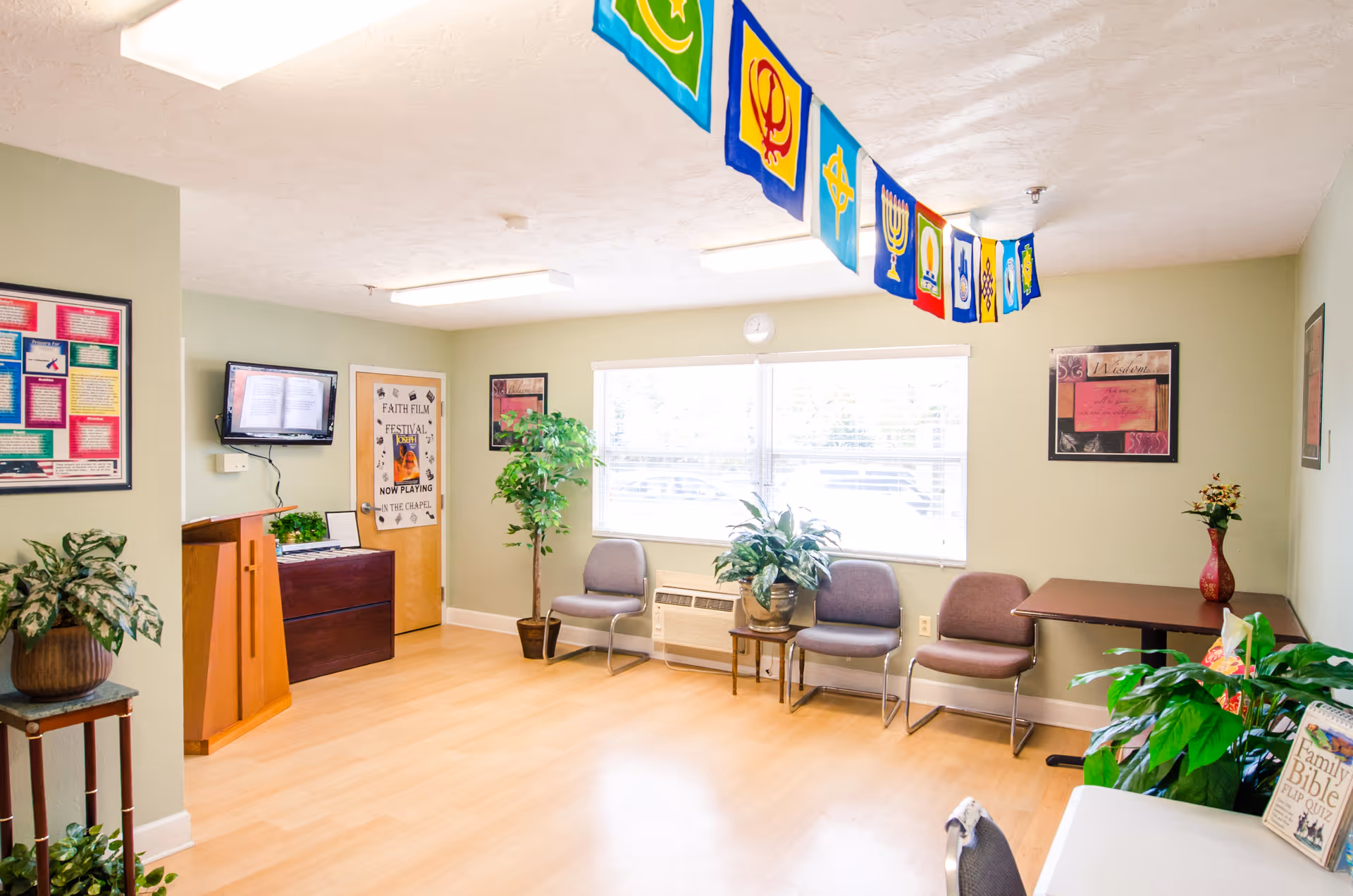 A bright, clean room with light green walls and wood flooring, featuring several chairs arranged along the walls, potted plants, a small table with a vase of flowers, a wall-mounted TV, and colorful religious flags hanging from the ceiling.