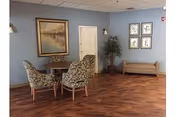 A cozy interior room with light blue walls and wood flooring featuring a small round table with four floral upholstered chairs. The walls are decorated with a large framed painting, four smaller framed artworks, and a tall potted plant. There is also a beige cushioned bench against the wall.