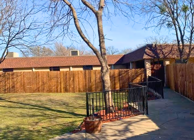 Outdoor area of a facility with a paved walkway, black metal railings, a tree in a planter, a wooden fence, and a building with a brown roof in the background under a clear blue sky.