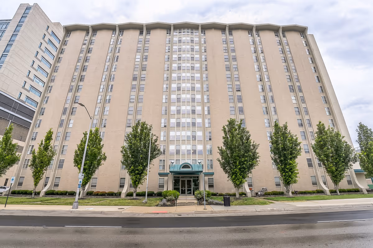 Tall beige multi-story building with rows of windows, a central entrance canopy, and trees along the sidewalk.