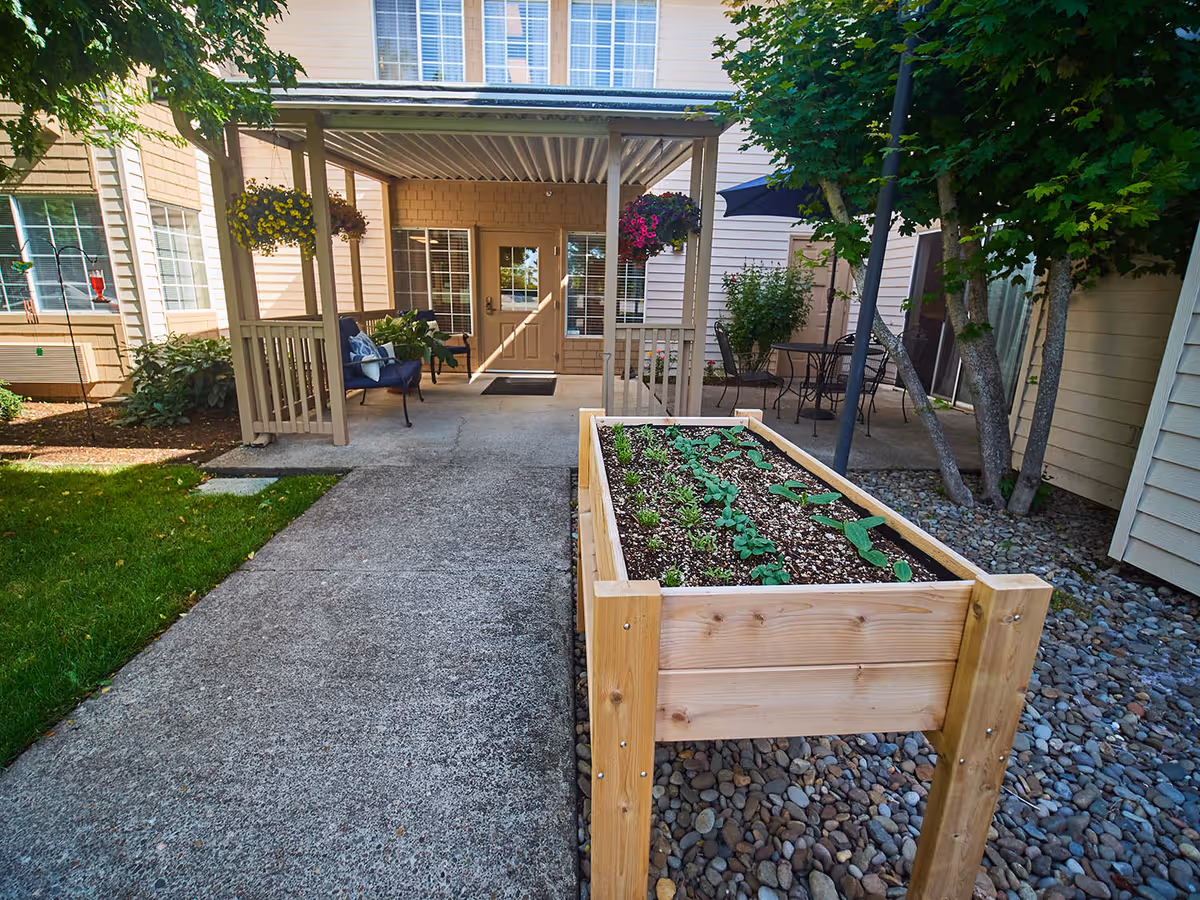 Outdoor patio area at Emerald Gardens featuring a raised wooden garden bed with young plants, a covered seating area with chairs and hanging flower baskets, and additional patio furniture under an umbrella surrounded by trees and shrubs.