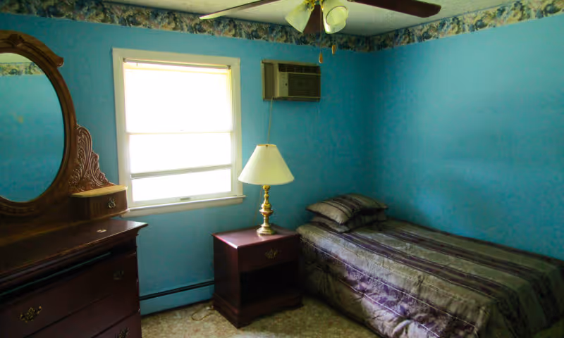 A small bedroom with blue walls and floral wallpaper border near the ceiling. The room contains a single bed with striped bedding, a wooden nightstand with a brass lamp, a wooden dresser with an ornate mirror, and a window with blinds. There is a ceiling fan with lights and a wall-mounted air conditioning unit.