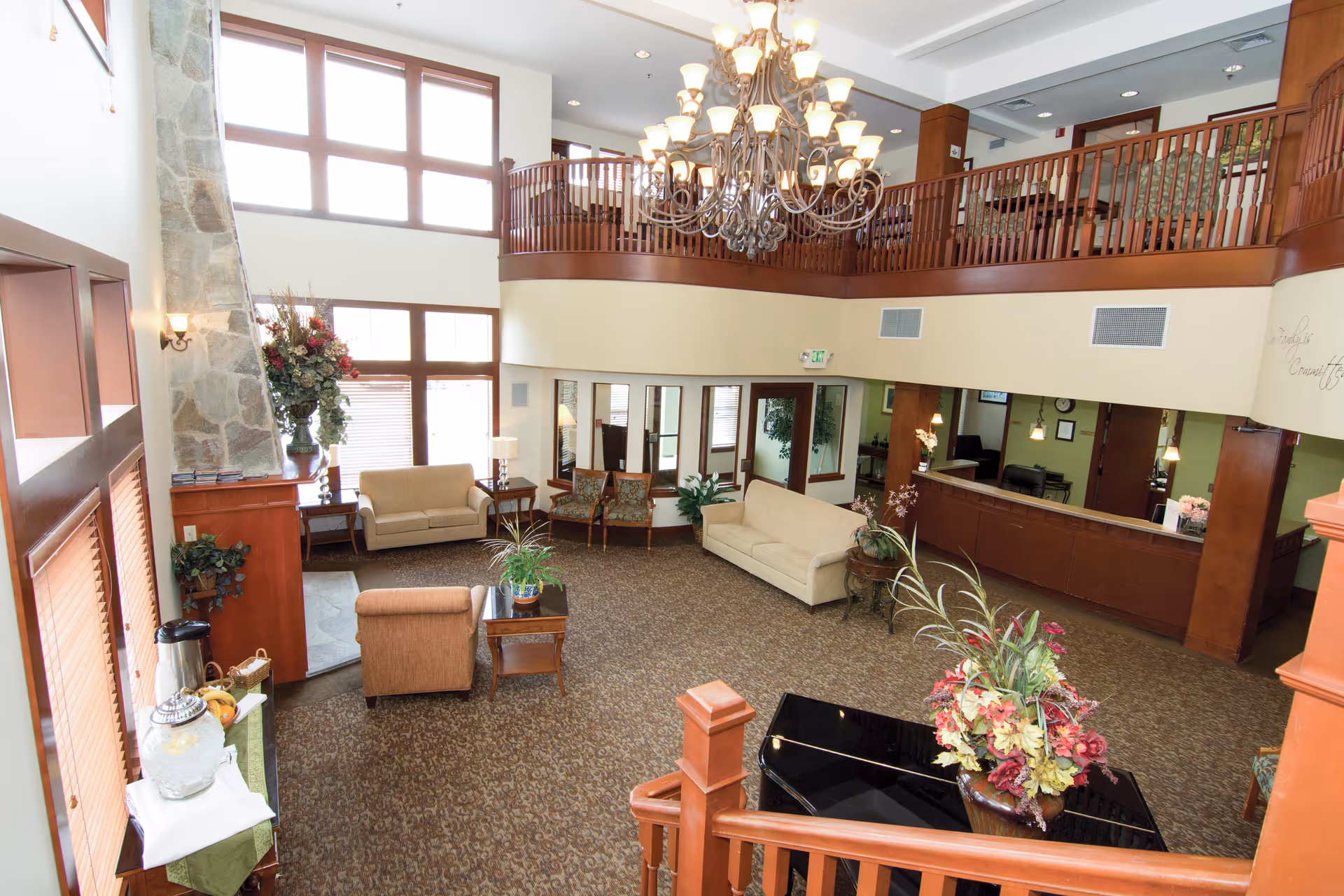 Spacious senior living facility lobby with high ceilings, large windows, a stone fireplace, multiple seating areas including sofas and armchairs, a chandelier, and a reception desk. There is a grand piano with a floral arrangement on top and a staircase with wooden railings leading to an upper balcony.