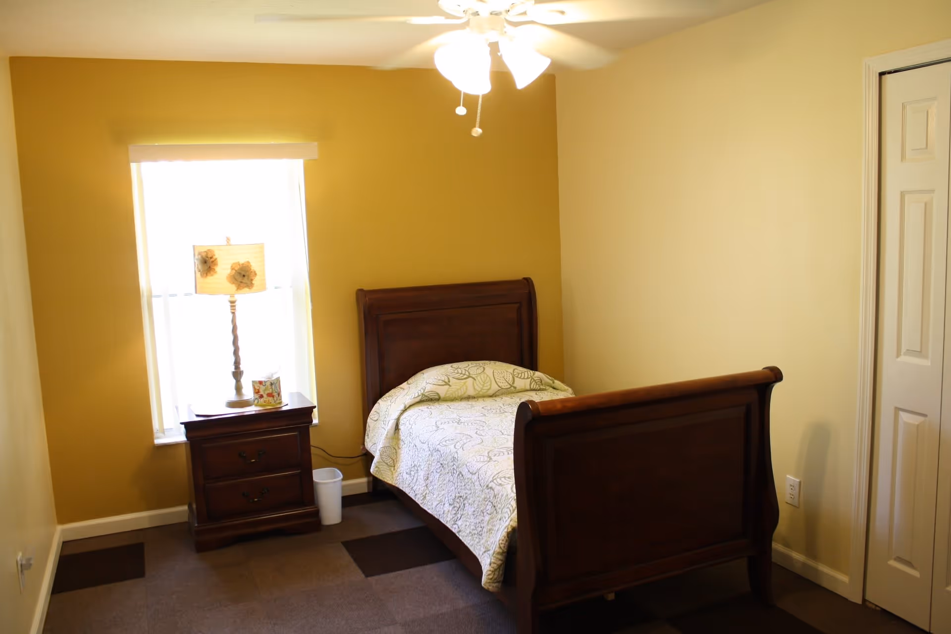A small bedroom with a single wooden bed featuring a patterned bedspread, a wooden nightstand with a decorative lamp and a small item on it, a window with blinds, a ceiling fan with lights, and a closed white door on the right side.