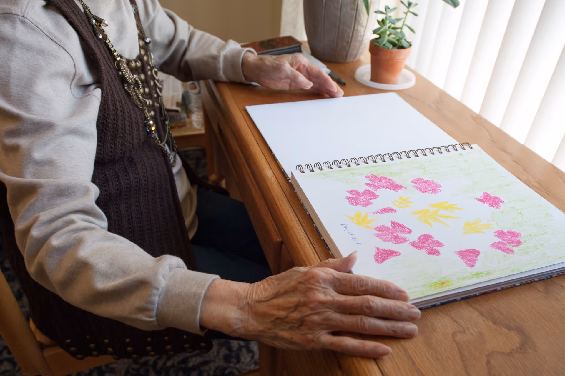 An elderly person sitting at a wooden table with a sketchbook open, displaying colorful crayon drawings of pink and yellow shapes. A small potted plant is on the table near a window with vertical blinds.