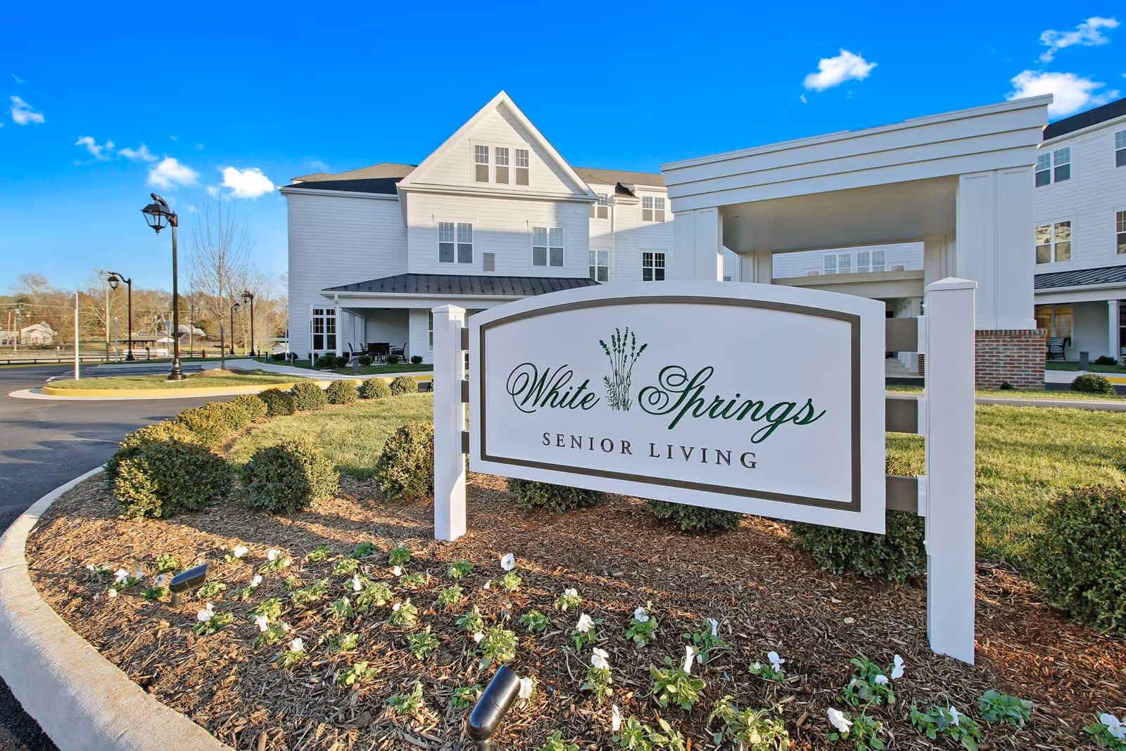 Exterior view of White Springs Senior Living facility with a clear blue sky, landscaped garden with small bushes and flowers, a paved driveway, and a large white sign displaying the facility's name in front of the building.