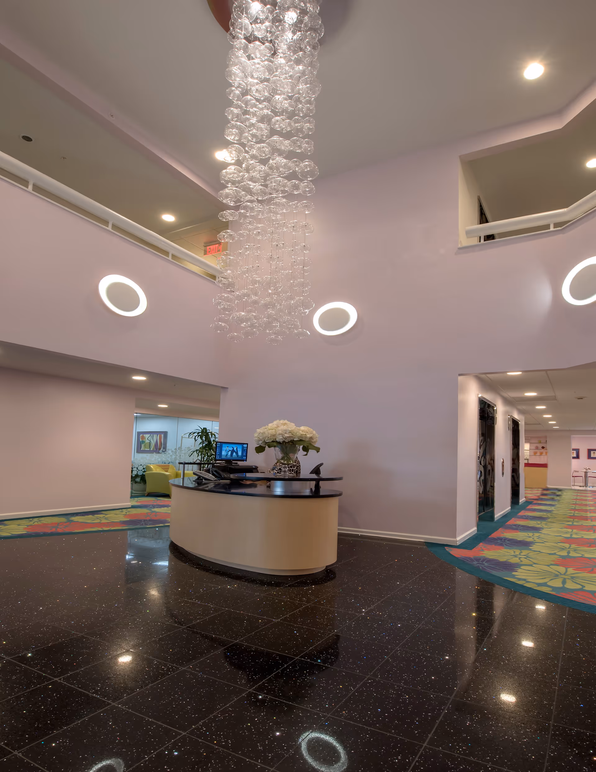 Interior view of a senior living facility lobby with a modern circular reception desk adorned with a vase of white flowers. The floor is shiny black tile, and the walls are light-colored with circular wall lights. A large, elegant chandelier made of multiple glass spheres hangs from the ceiling. There is a colorful carpeted hallway leading to other rooms.