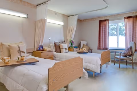 A bright and clean rehabilitation room with two hospital beds, each equipped with white bedding and pillows. There are bedside tables with flowers and personal items, a window with red curtains letting in natural light, and a chair near the window. Privacy curtains hang between the beds.