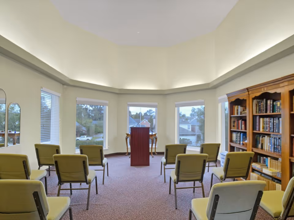 A small meeting or presentation room with green cushioned chairs arranged in rows facing a wooden podium. The room has large windows with white blinds, allowing natural light to enter. There is a wooden bookshelf filled with books on the right side of the room. The walls are painted light yellow, and the ceiling has recessed lighting.