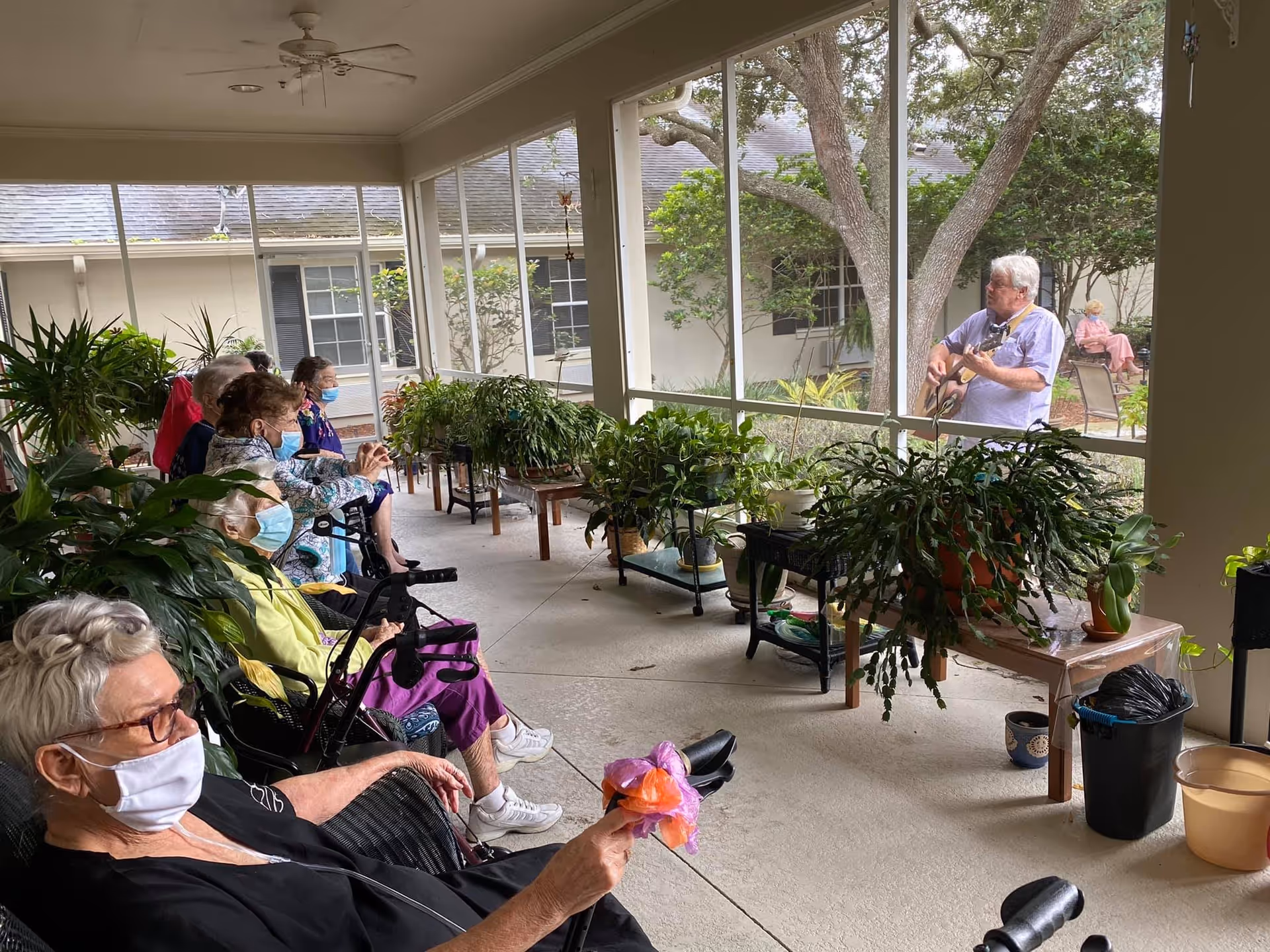 A group of elderly people wearing face masks sitting on chairs and wheelchairs on a covered patio, watching a man playing guitar outside the screened area. The patio is decorated with various potted plants on tables and stands.