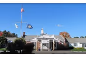 Front exterior view of Webster at Rye facility with a flagpole displaying the American flag and another flag, a brick building with white trim, and a clear blue sky.