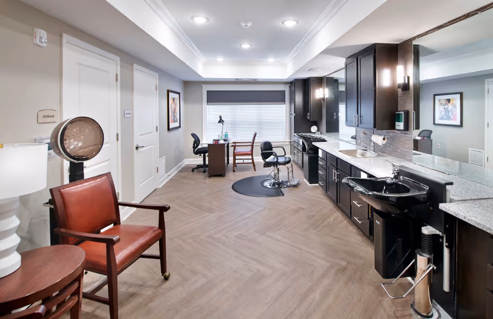 Interior view of a salon area in a senior living facility with a hair dryer chair on the left, a styling chair in the center on a black mat, and a long counter with sinks and cabinets on the right. There is a desk with two chairs near a window with blinds at the back of the room.