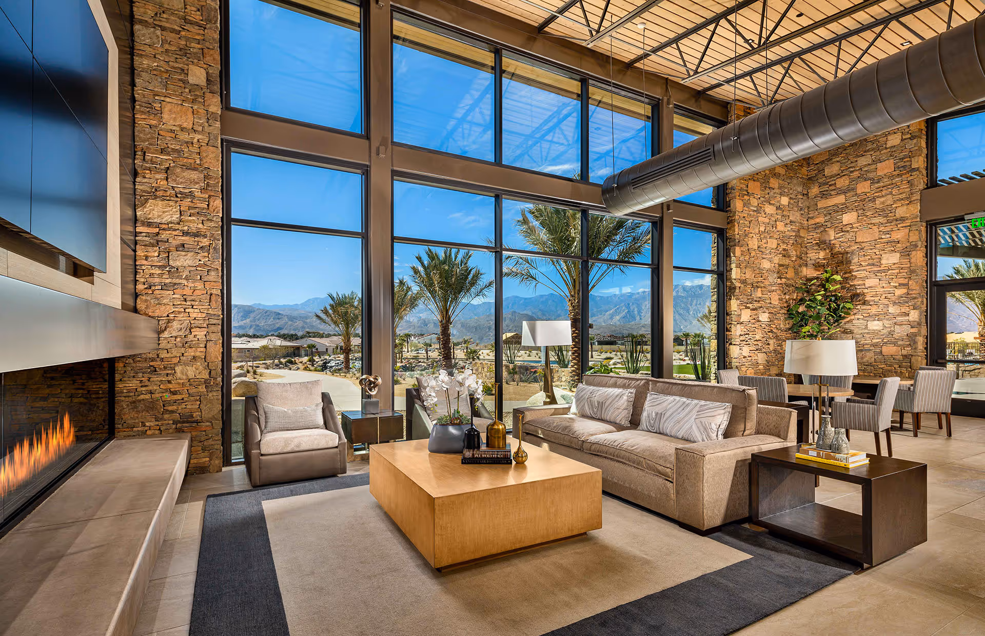 A spacious and modern living room with large floor-to-ceiling windows offering a view of palm trees and mountains outside. The room features a stone accent wall with a built-in fireplace, a beige sofa with patterned pillows, a matching armchair, a wooden coffee table with decorative items, and side tables with lamps. The ceiling has exposed ductwork and a wooden slatted design.