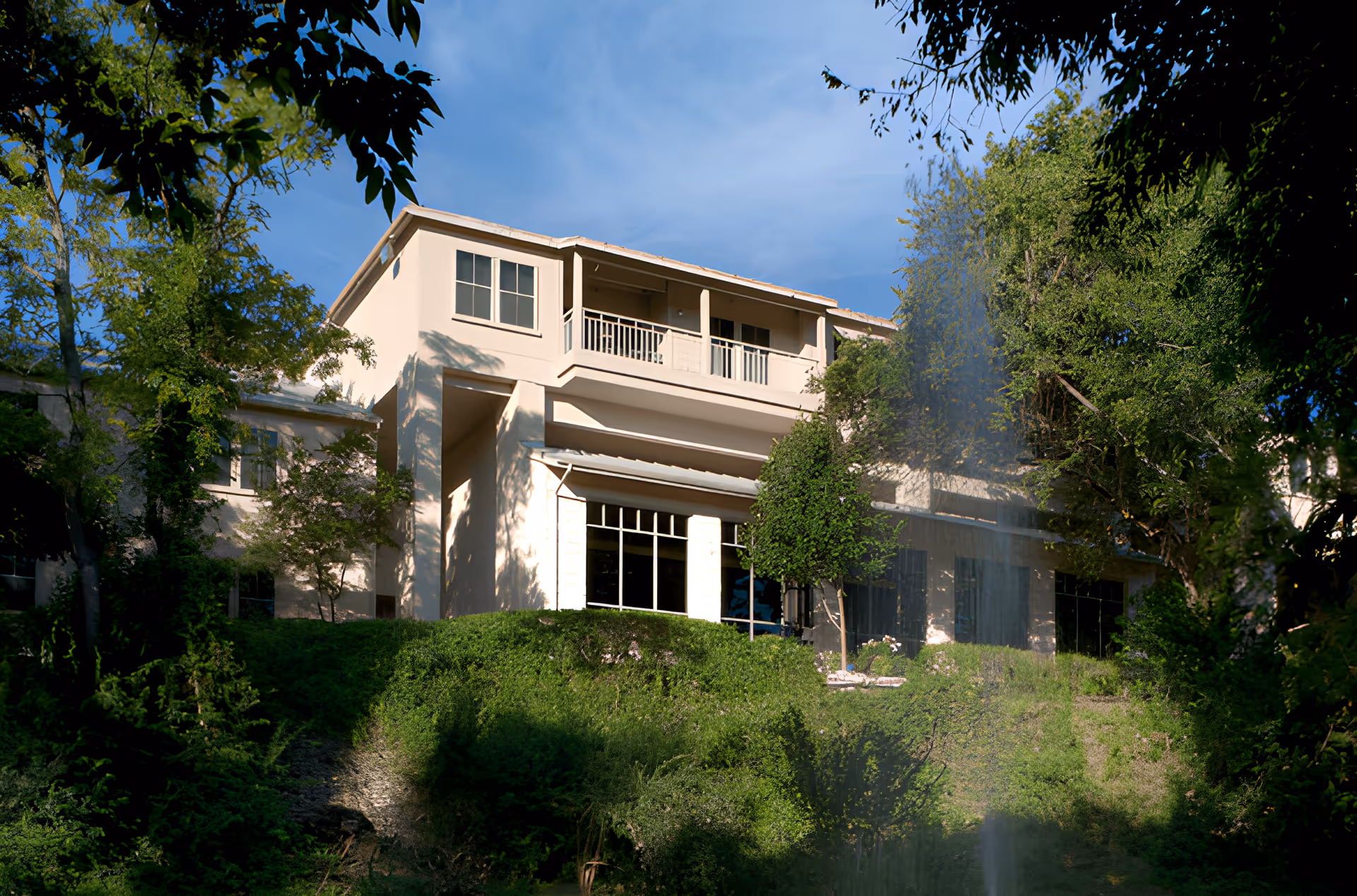 Exterior view of a two-story beige building surrounded by lush green trees and bushes under a clear blue sky. The building has large windows and a balcony on the upper floor. A water fountain is visible in the foreground.