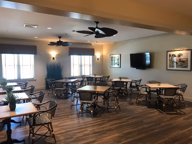 A dining room with several square wooden tables and black and white woven chairs arranged neatly. The room has large windows with blinds, two ceiling fans, wall-mounted lights, framed artwork, and a flat-screen TV mounted on the wall. The floor is wooden, and there are some decorative plants near the windows.