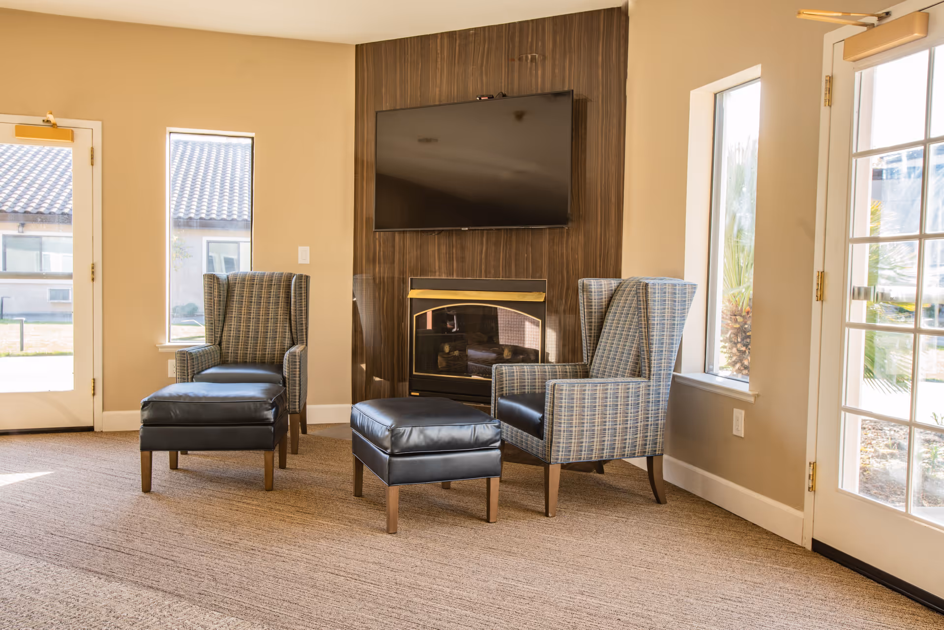 A cozy seating area with two patterned armchairs and two matching black leather ottomans positioned in front of a wall-mounted flat-screen TV and a fireplace. The room has beige walls, carpeted floor, and large windows and glass doors letting in natural light.