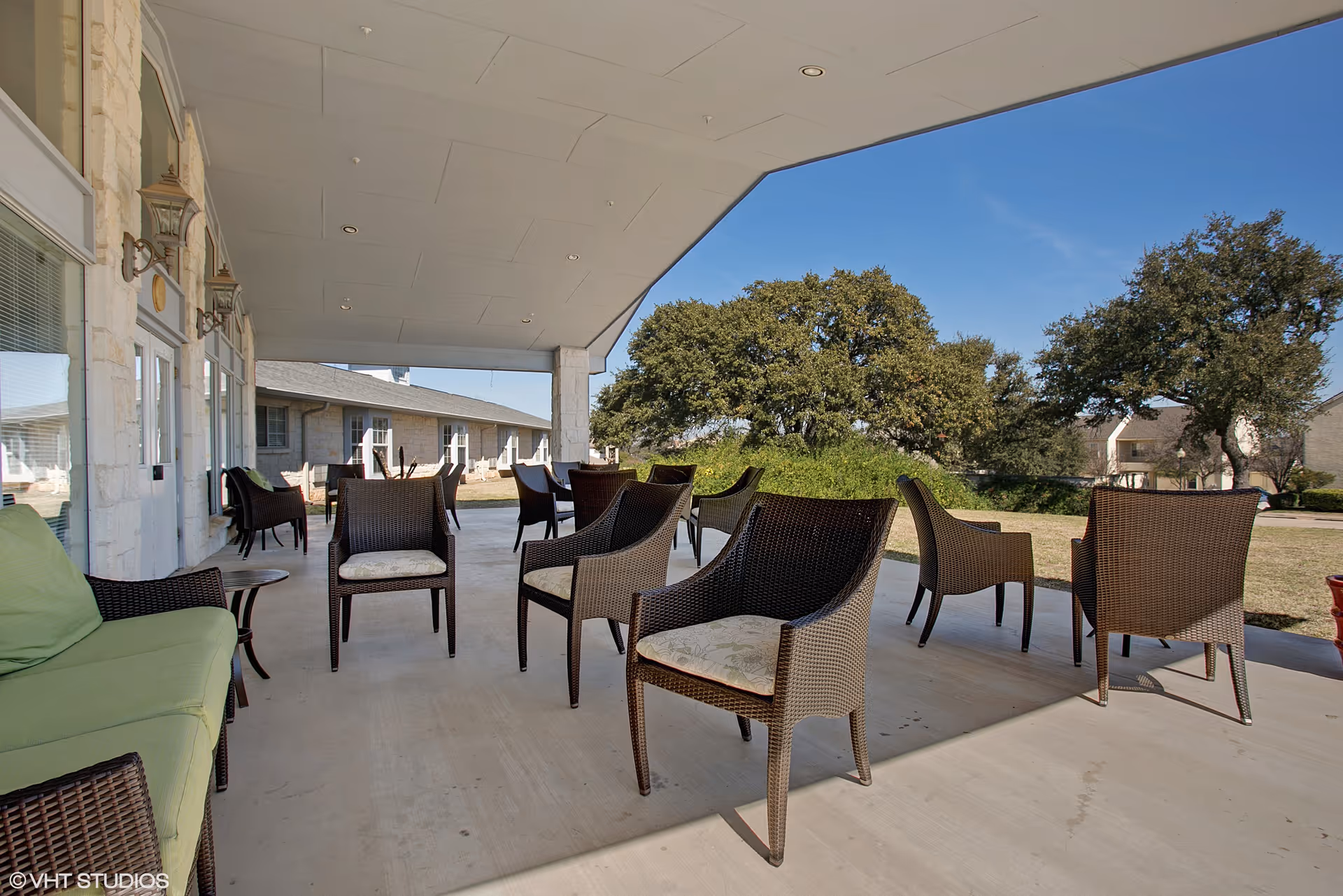 Covered outdoor patio area with multiple wicker chairs and a green cushioned sofa. The patio overlooks a grassy area with trees and other buildings in the background under a clear blue sky.