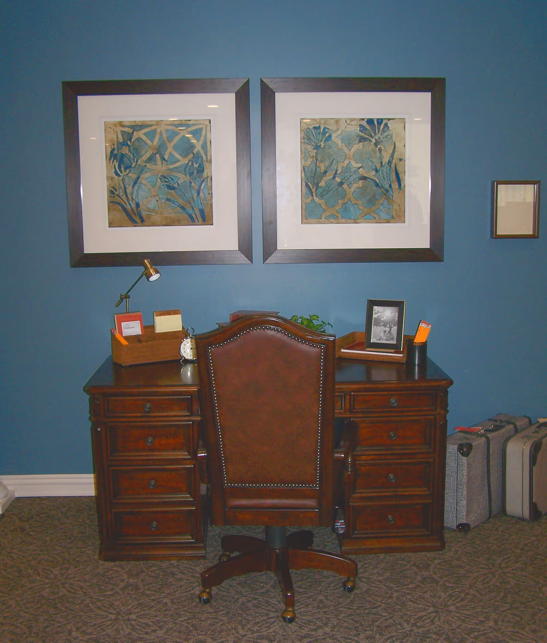 A traditional wooden desk with multiple drawers and a brown leather office chair in front of it. On the desk are a small lamp, an alarm clock, a photo frame, a pencil holder with pencils, and some small decorative items. Two framed abstract floral artworks hang on the blue wall above the desk. Two suitcases are placed on the floor to the right of the desk.