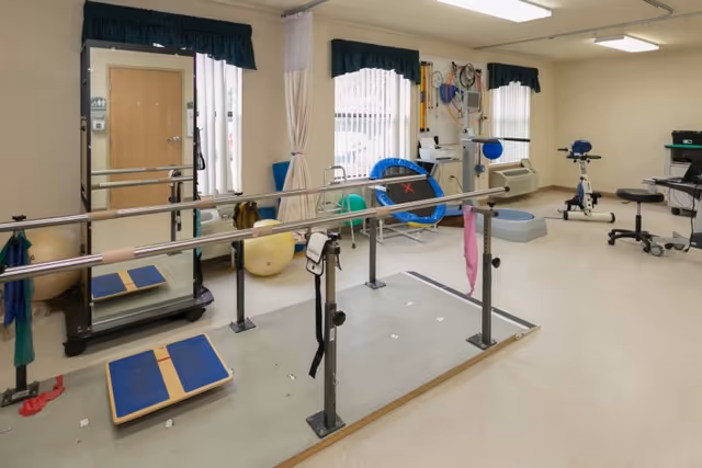 Physical therapy room with parallel bars for walking exercises, balance boards, exercise balls, a small trampoline, stationary bike, and various therapy equipment. The room has beige walls, windows with dark green valances, and a door on the left side.
