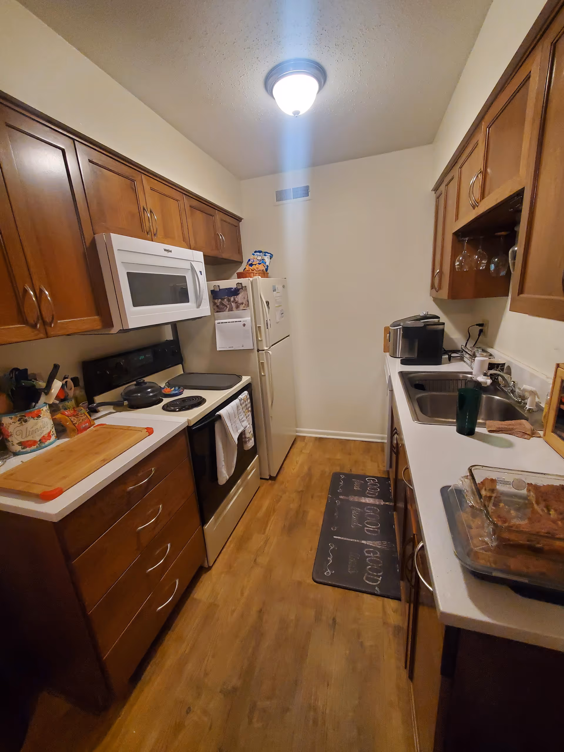 A narrow kitchen with wooden cabinets on both sides, a white microwave above a stove with pots, a refrigerator at the end, and a countertop with a sink, coffee maker, and a glass baking dish. The floor is wooden, and a ceiling light illuminates the space.
