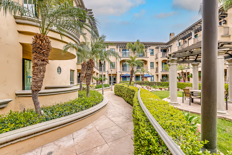 Landscaped central courtyard with a paved walkway, palm trees, hedges, seating areas and surrounding Mediterranean-style building.