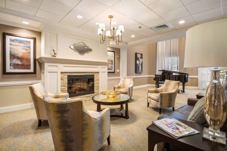 A cozy living room area with four upholstered armchairs arranged around a round glass coffee table. The room features a decorative fireplace with a mirror above it, framed artwork on the walls, a chandelier hanging from the ceiling, and a grand piano near large windows with sheer curtains. A side table with a lamp and magazines is also visible.