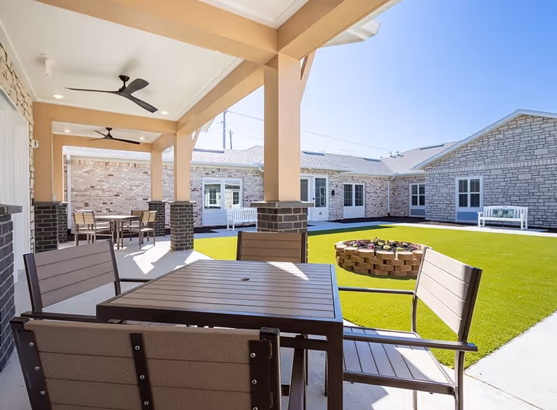 Outdoor covered patio area with ceiling fans, brown wooden tables and chairs, overlooking a green lawn with a circular brick planter and benches, surrounded by light-colored brick buildings under a clear blue sky.