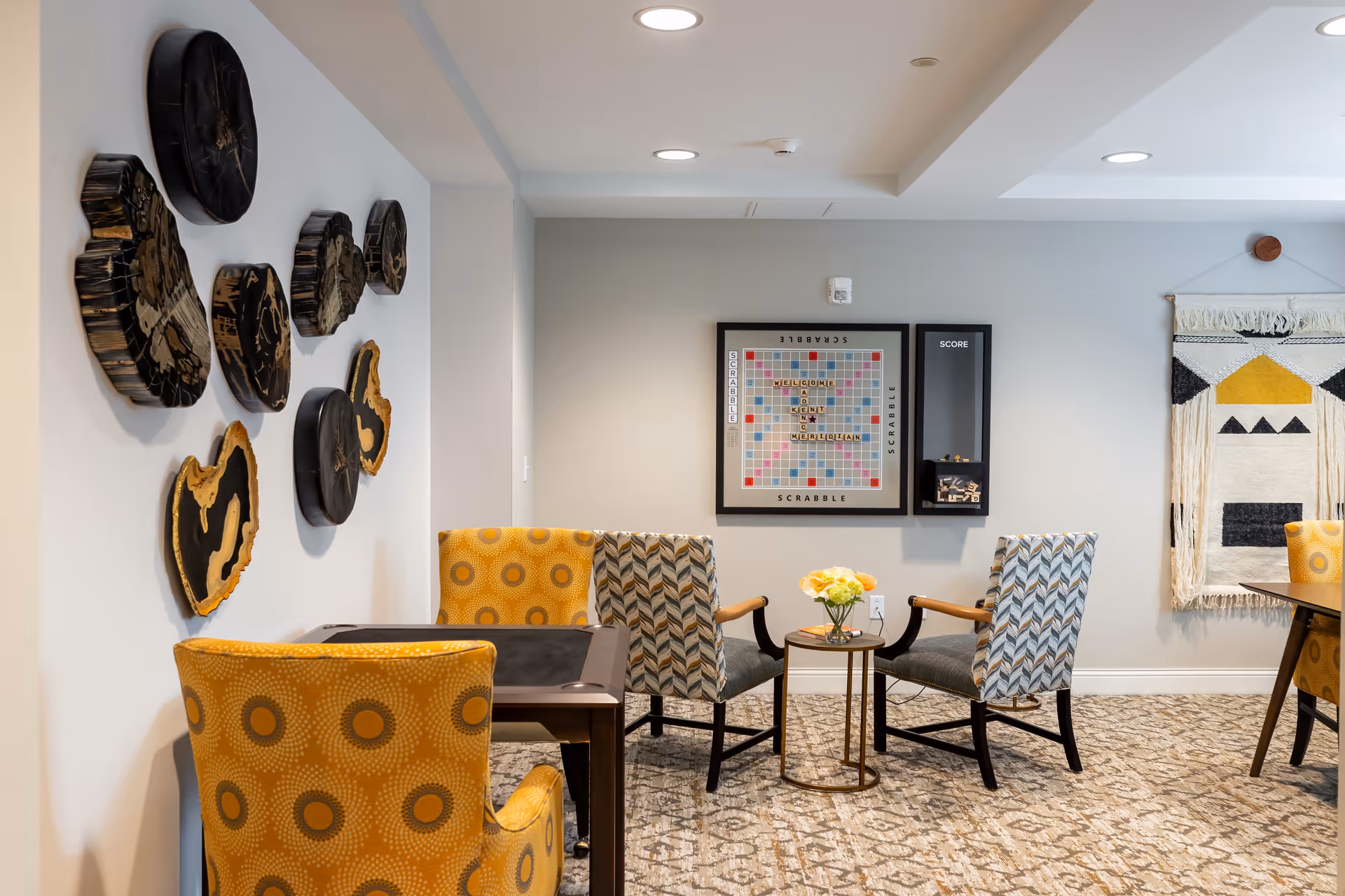 A cozy interior common area with patterned chairs arranged around small tables. On the wall is a framed Scrabble board with the words 'WELCOME TO KENT MERIDIAN' spelled out, and decorative wooden wall art on the adjacent wall. The room has soft lighting and a patterned carpet.