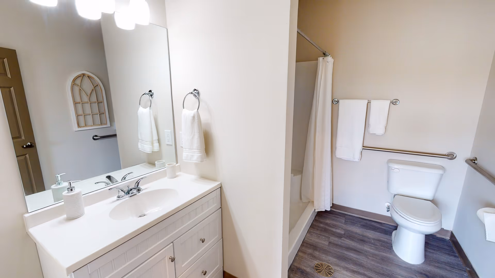 A clean and accessible bathroom with a white vanity and sink, a large mirror above the sink, a towel ring with a white towel, a shower with a white curtain, a toilet with grab bars on the walls, and wood-look flooring.