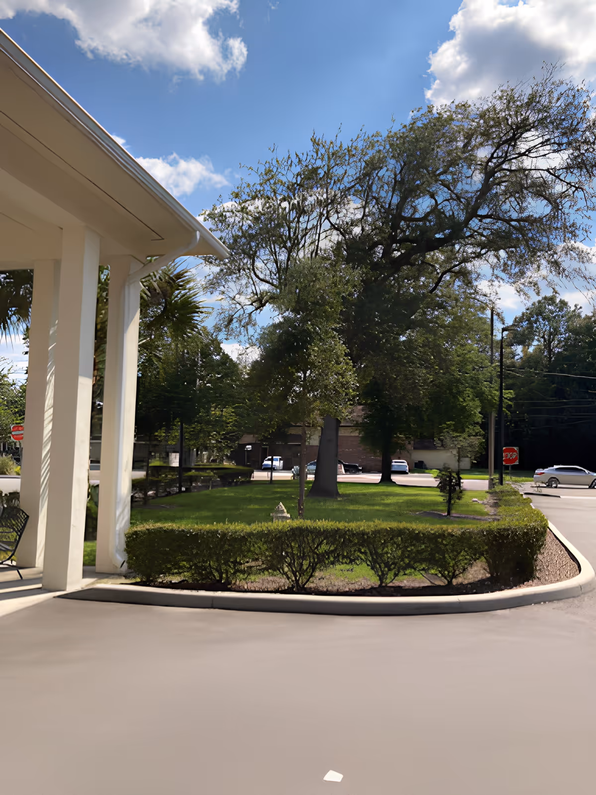 View of the outdoor area at Noble House of Jacksonville showing a covered driveway with white pillars, neatly trimmed hedges, green grass, large trees, and a partly cloudy blue sky. Several parked cars and a stop sign are visible in the background.