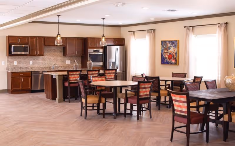 A spacious dining area with multiple round and rectangular tables surrounded by chairs with red and beige checkered upholstery. In the background, there is a kitchen area with dark wooden cabinets, stainless steel appliances including a refrigerator, oven, microwave, and dishwasher. Two pendant lights hang over a kitchen island with three bar stools. The room has light-colored walls, large windows with beige curtains, and a colorful framed painting on the wall.