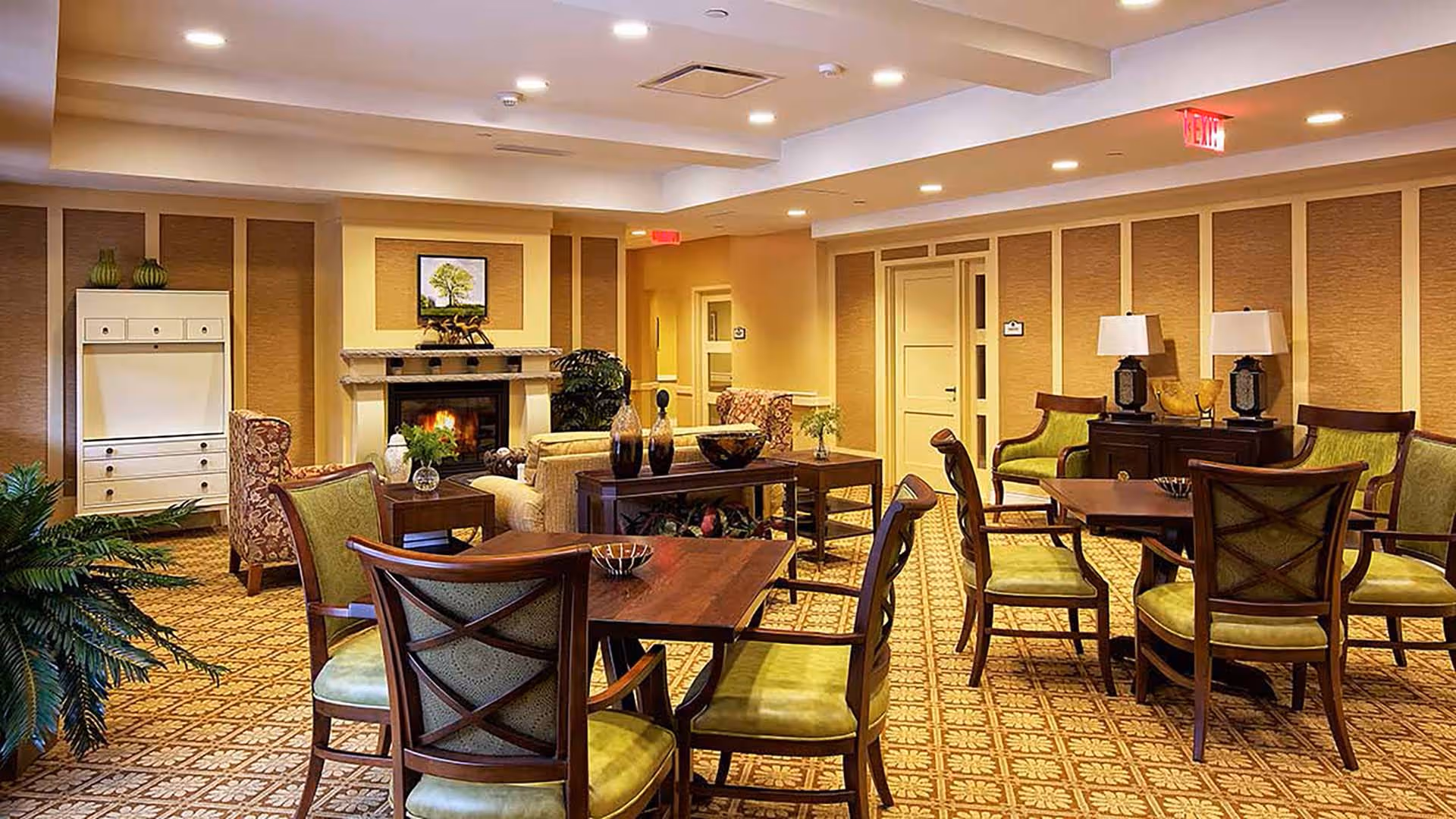 A warm and inviting senior living facility common area with multiple wooden tables and green cushioned chairs arranged for socializing. The room features a cozy fireplace with a framed picture above it, decorative vases on tables, patterned carpet, and soft lighting from ceiling fixtures. There are also armchairs and plants adding to the comfortable ambiance.